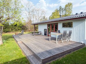 Una terraza de madera con una mesa de picnic y sillas, un área de estar cubierta y un pequeño edificio con una ventana.