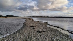 A coastal landscape with a pebbly beach, calm water, and a distant shoreline.