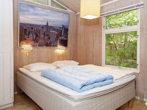 A bedroom with a bed, a cityscape painting, two wall-mounted lights, and a window with a view of greenery.