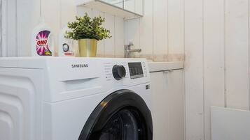 A white Samsung washing machine in a utility room with a shelf above it holding a bottle of OMO detergent and a potted plant.