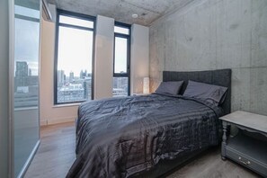 Un dormitorio con una cama grande, una mesita de noche y vistas a un paisaje urbano a través de amplias ventanas.