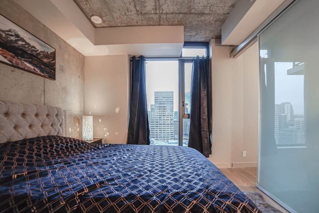A bedroom with a large bed, a tufted headboard, a city view through a window, and a modern ceiling.