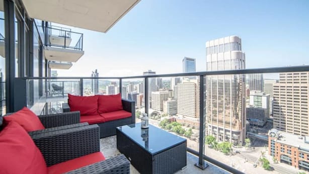 Un balcón con muebles de mimbre y cojines rojos, con vista a un paisaje urbano con edificios modernos.