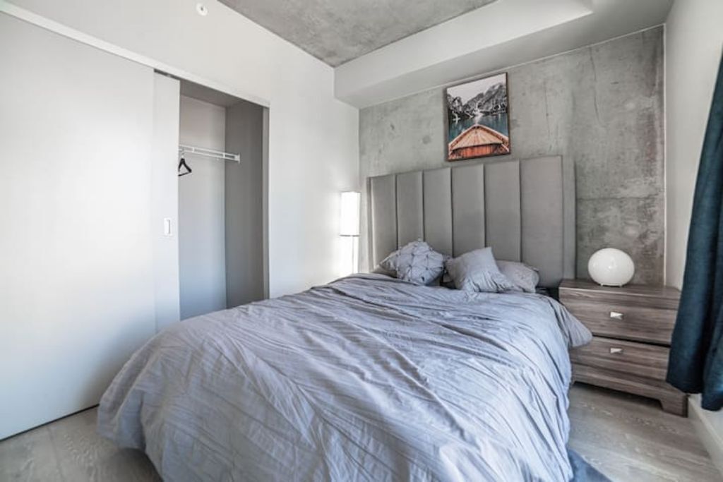 Une chambre à coucher moderne avec un lit gris, une tête de lit à lattes verticales, un tableau au mur et une table de chevet avec une lampe.