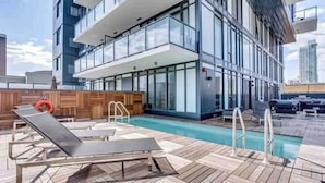 Un área de piscina en la azotea con tumbonas, una terraza de madera y un edificio de vidrio al fondo.