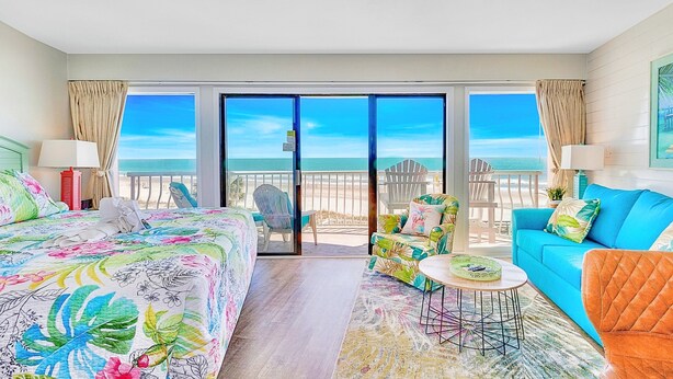 Ein Hotelzimmer mit einem großen Bett, einem blauen Sofa, einem kleinen Tisch und Blick auf den Strand durch eine Schiebetür.