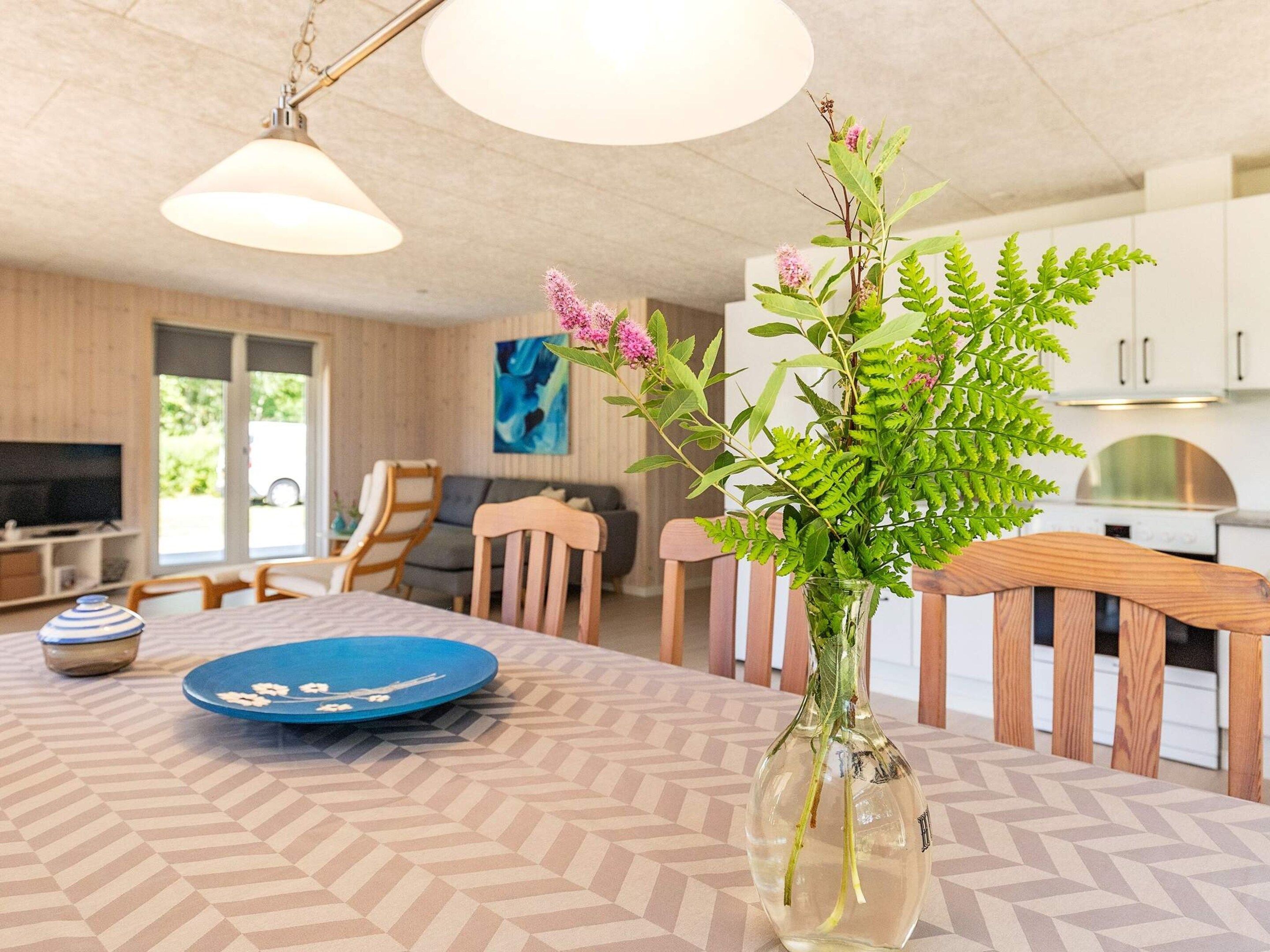 Uma cozinha moderna com armários brancos, uma mesa de jantar com toalha de mesa estampada e um vaso com folhagem verde e flores cor-de-rosa.