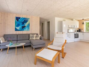 Ein modernes Wohnzimmer mit einem grauen Ecksofa, einem kleinen runden Tisch und einem Holzsesseln. Ein großes abstraktes Gemälde ziert die Wand, und der Raum geht in eine Küche mit weißen Schränken und einem Herd über.