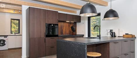 A modern kitchen with wooden cabinets, a marble island, pendant lighting, and a view of trees through the window.