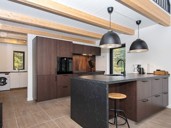 Una cocina moderna con armarios de madera, una isla de mármol, iluminación colgante y vistas a árboles por la ventana.