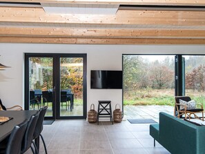 A modern living room with a large sliding glass door leading to a garden, a flat-screen TV mounted on the wall, and a dining area with a table and chairs.