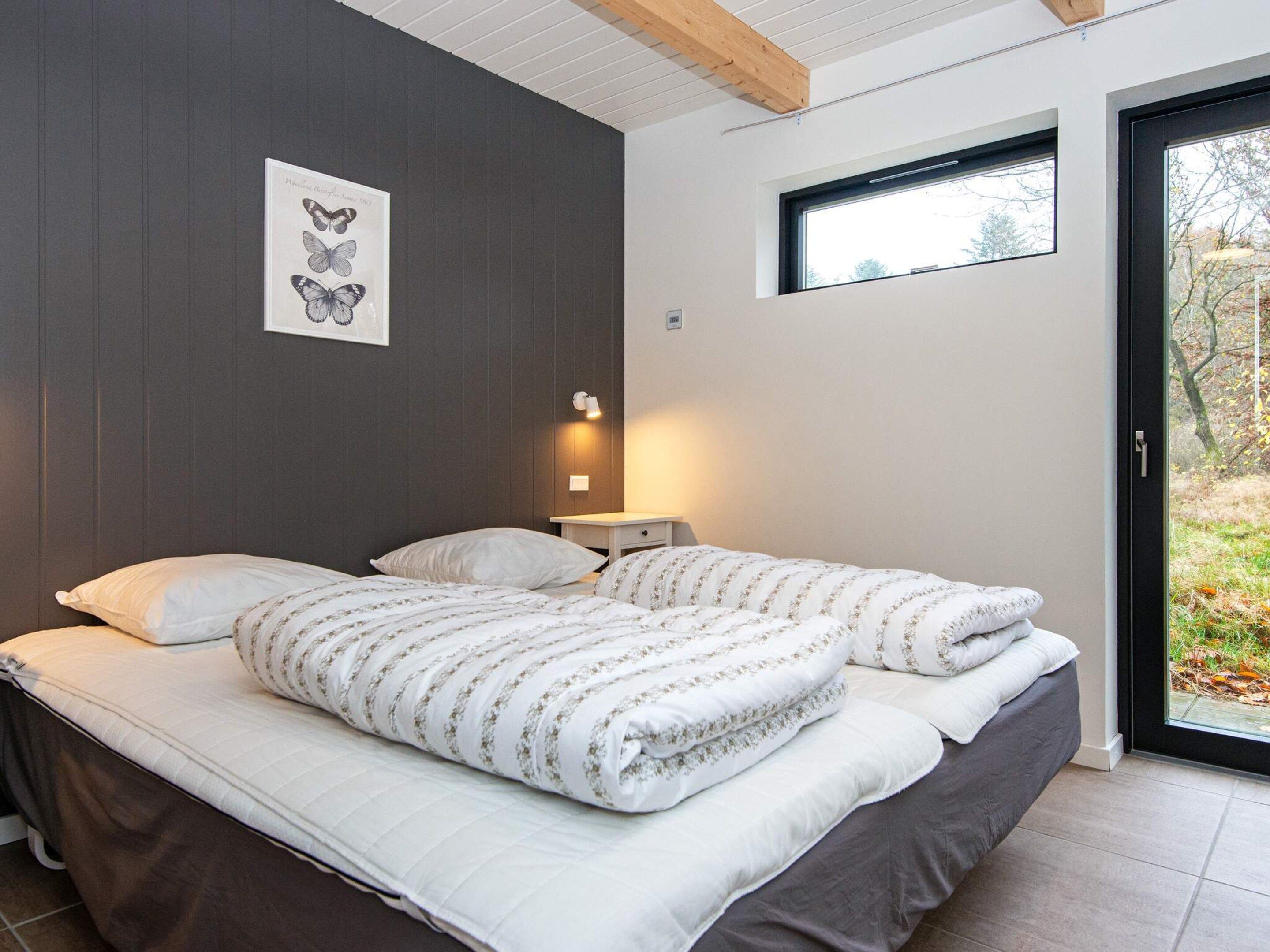 A bedroom with a bed, pillows, a bedside table with a lamp, a framed botanical illustration on the wall, and a window with a view of trees.