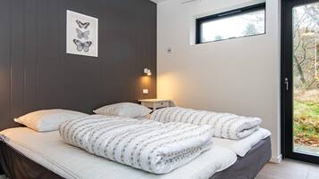 A bedroom with a bed, pillows, a bedside table with a lamp, a framed botanical illustration on the wall, and a window with a view of trees.