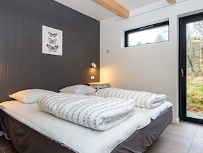 A bedroom with a bed, pillows, a bedside table with a lamp, a framed botanical illustration on the wall, and a window with a view of trees. - 6 Person Holiday Home in Hovborg (Hovborg)