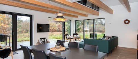 A modern dining area with a large table and chairs, a television mounted on the wall, and a view of the outdoors through large windows.