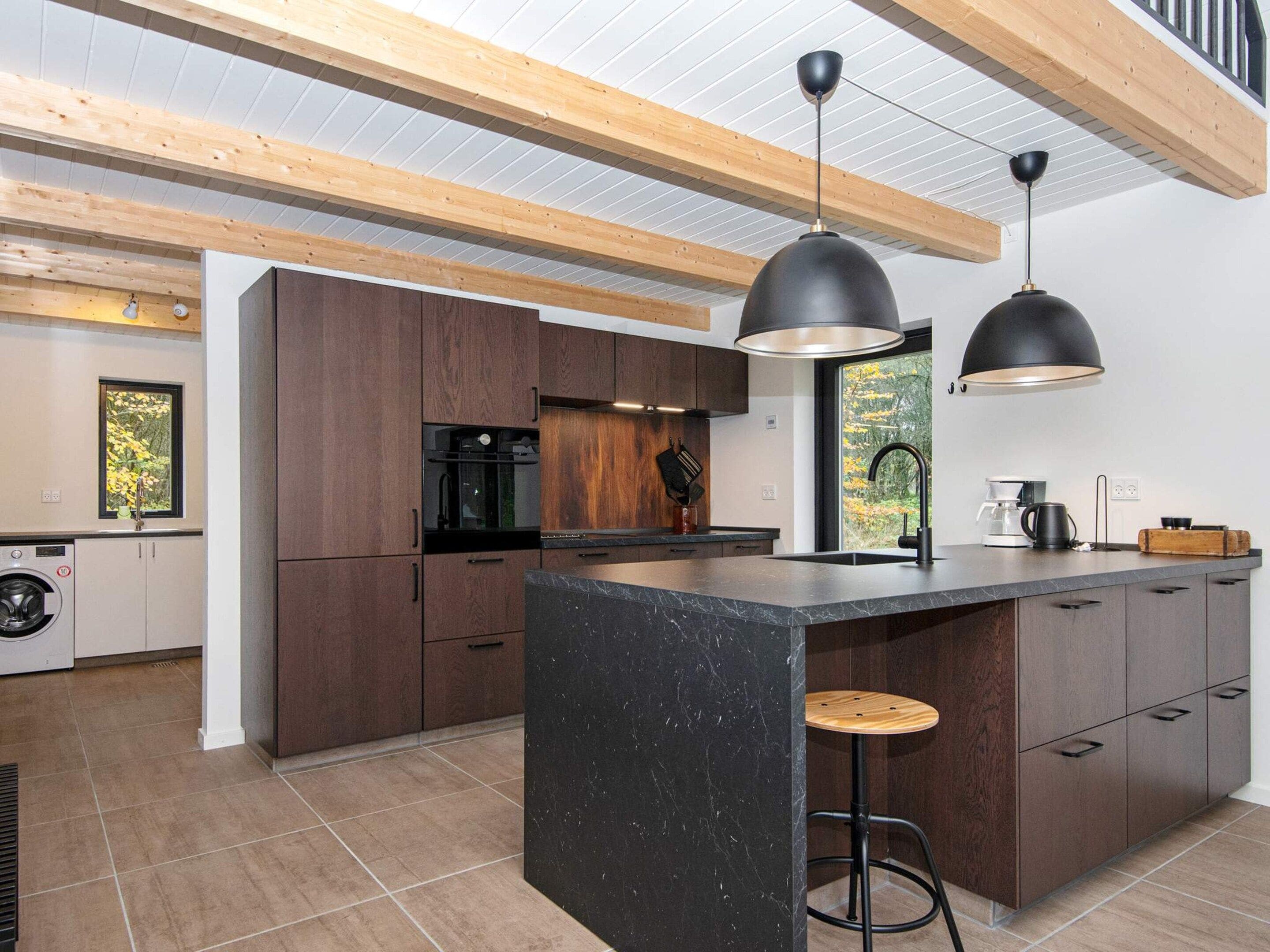 Una cocina moderna con armarios de madera, una isla de mármol, iluminación colgante y vista a los árboles por una ventana.