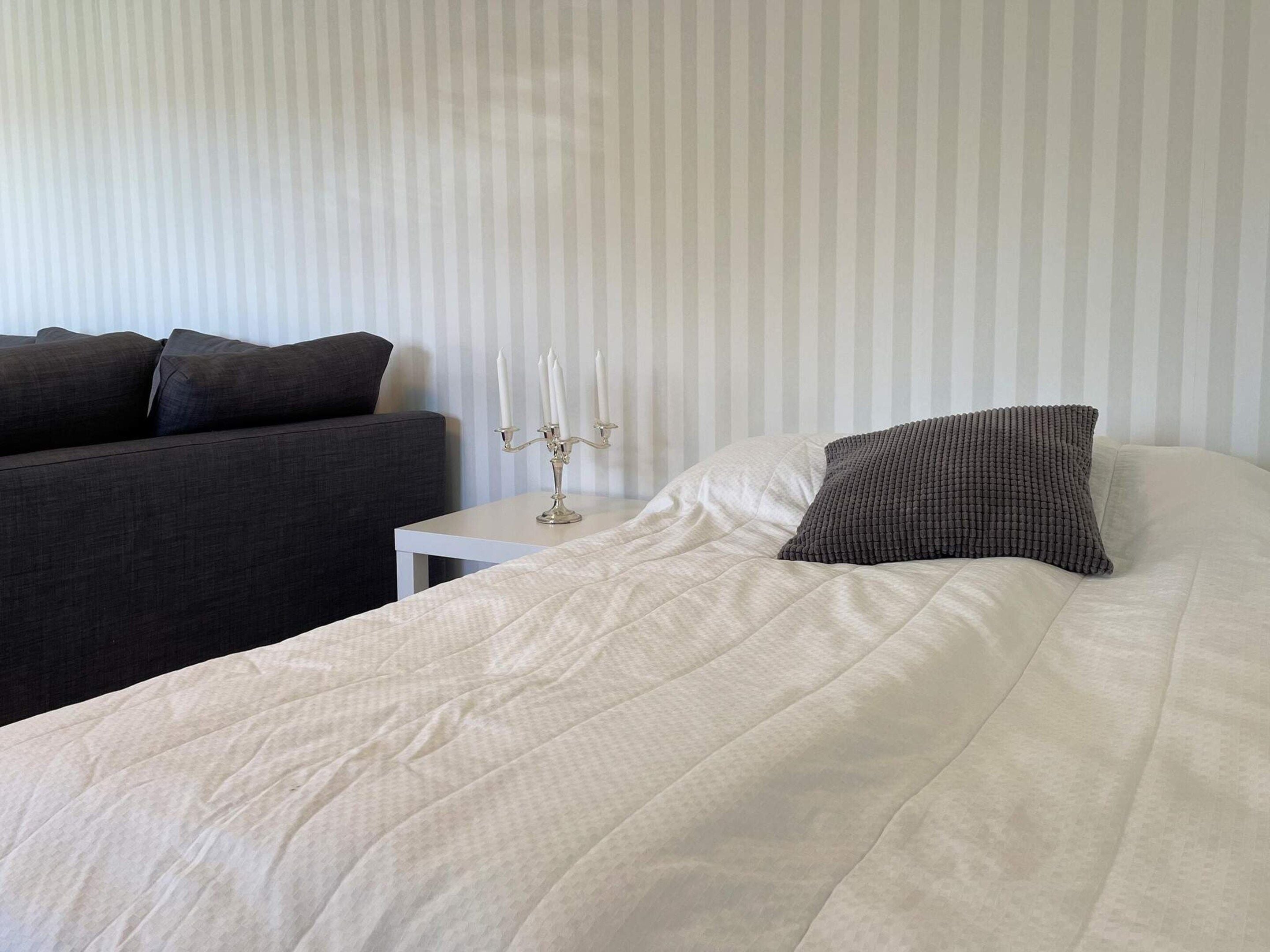 Una cama bien tendida con edredón blanco y almohada texturizada, una mesita de noche con candelabro y un sofá oscuro frente a una pared a rayas.