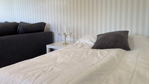 Ein ordentlich bezogenes Bett mit weißer Bettdecke und einem strukturierten Kissen, ein Nachttisch mit einem Kerzenleuchter und ein dunkles Sofa an einer gestreiften Wand.