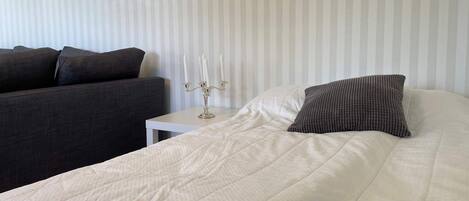 A neatly made bed with a white duvet and a textured pillow, a bedside table with a candelabra, and a dark sofa against a striped wall.