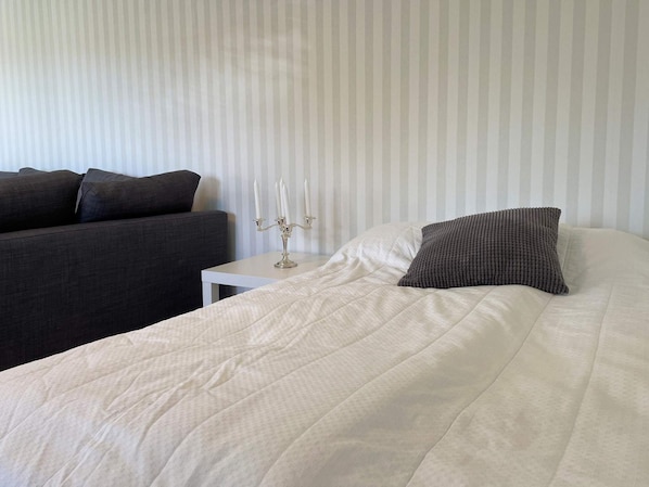 A neatly made bed with a white duvet and a textured pillow, a bedside table with a candelabra, and a dark sofa against a striped wall. - 4 Star Holiday Home in Skarhamn (Skaerhamn)