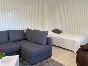 A modern living room with a grey sectional sofa, a single bed with a white duvet, and a small table with a vase of flowers. - 4 Star Holiday Home in Skarhamn (Skaerhamn)