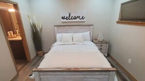 A bedroom with a wooden bed frame, a 'welcome' sign above the bed, a nightstand with a lamp, and a potted plant. - Beautiful Cabin in Unbeatable Location (Pinetop-Lakeside)
