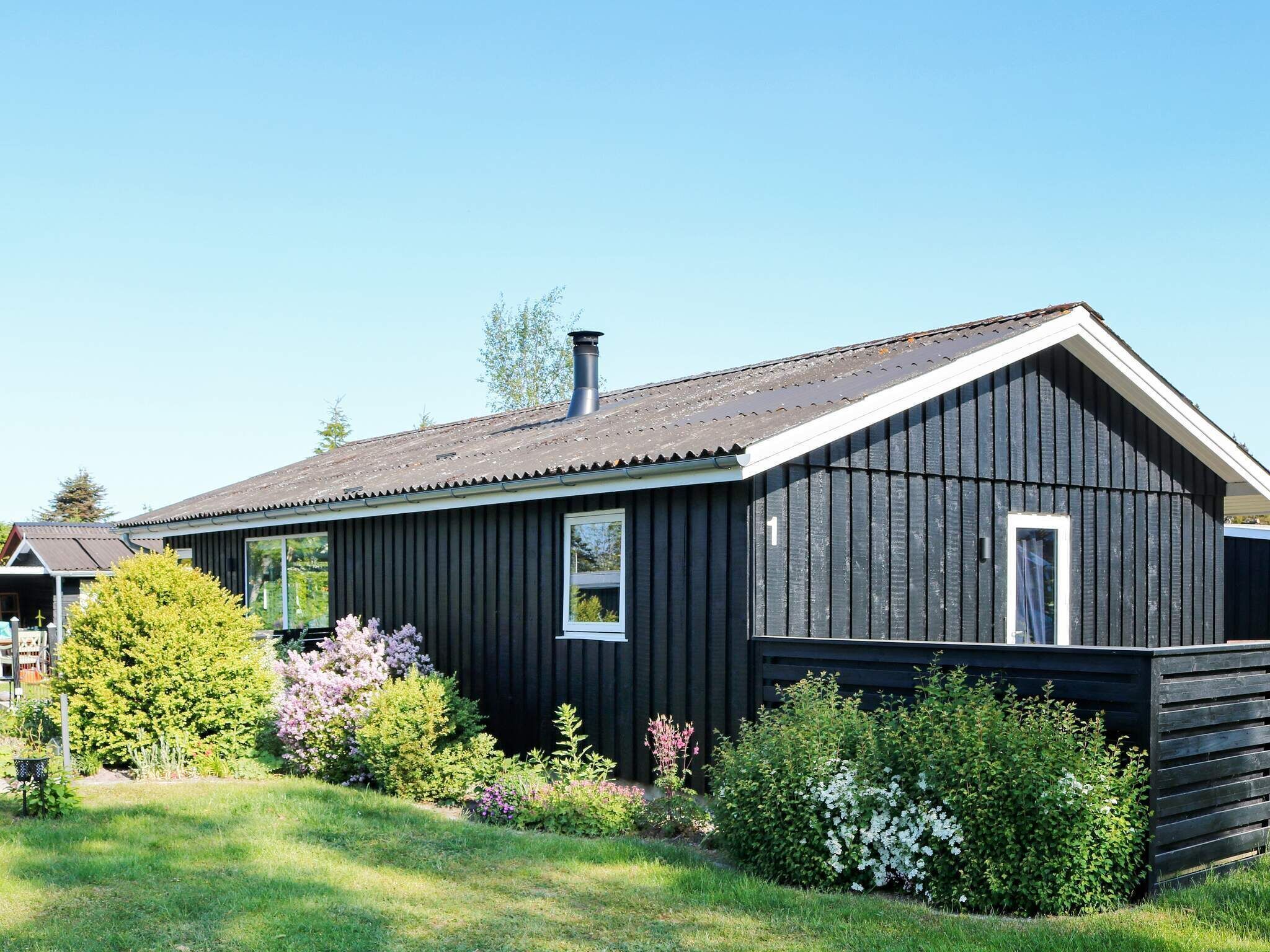 Une maison en bois noire, dotée d’un toit à pignon et d’une cheminée, entourée d’un jardin bien entretenu où poussent différentes espèces d’arbustes et de fleurs.