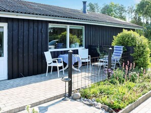 A black building with a tiled roof, a patio with white chairs and a table, and a garden with various plants. - 4 Person Holiday Home in Storvorde (Storvorde)
