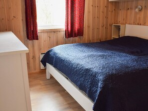 A bedroom with a wooden paneled wall, a white bedside table, a bed with a blue quilt, and red curtains. - 7 Person Holiday Home in Nesbyen (Nes)