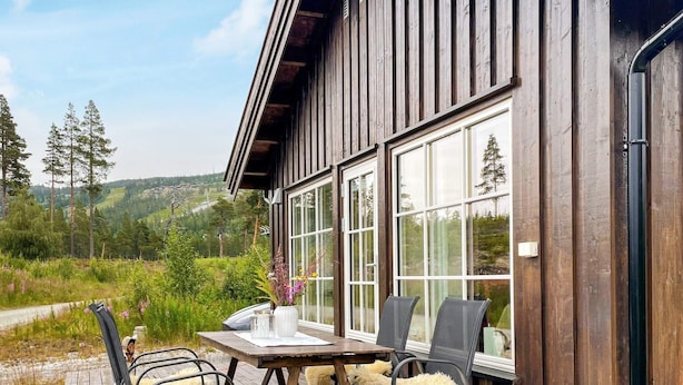 A wooden cabin with a deck, large windows, and a table with chairs.