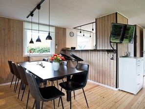 A modern kitchen with wooden paneling, a dining table with chairs, a TV mounted on the wall, and a window with blinds.