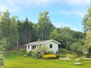 A small, single-story house with a sloped roof, surrounded by a lush green landscape and dense forest.