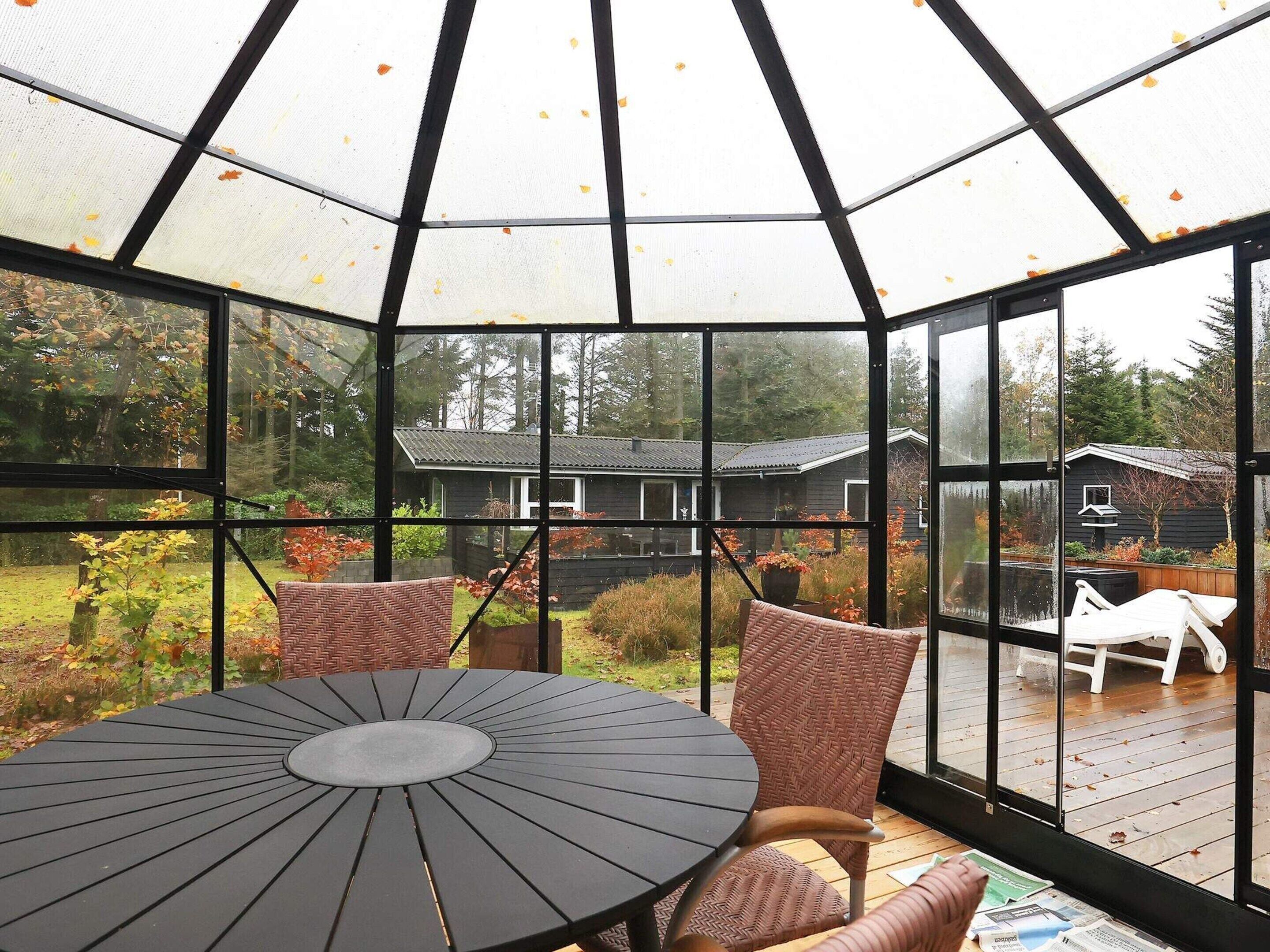 A conservatory with a round table and wicker chairs, offering views of a wooden house and garden.