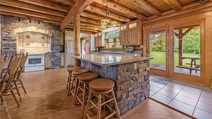 A rustic kitchen with a stone fireplace, wooden ceiling beams, and a bar area with stools. - Luxury Cabin w/Heated Pool, 22min to 7 Springs, HotTub, Sauna, Game Room & Views (Markleton)