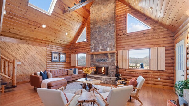 A log cabin interior with a stone fireplace, wooden furniture, and large windows. - Luxury Cabin w/Heated Pool, 22min to 7 Springs, HotTub, Sauna, Game Room & Views (Markleton)