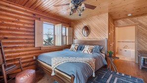 A log cabin bedroom with a bed, a ceiling fan, a window, a mirror, and a bathroom visible through an open door. - Luxury Cabin w/Heated Pool, 22min to 7 Springs, HotTub, Sauna, Game Room & Views (Markleton)