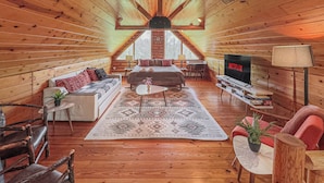 A cozy log cabin interior with a sofa, a bed, a TV, and a dining area. - Luxury Cabin w/Heated Pool, 22min to 7 Springs, HotTub, Sauna, Game Room & Views (Markleton)