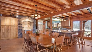A spacious kitchen with wooden cabinets, a stone fireplace, and a large dining table surrounded by chairs. - Luxury Cabin w/Heated Pool, 22min to 7 Springs, HotTub, Sauna, Game Room & Views (Markleton)