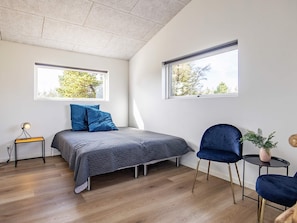 Een moderne slaapkamer met een bed, twee ramen, een stoel, een klein tafeltje en een plant.