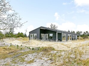 A modern, single-story building with large glass windows and a flat roof, situated on a sandy terrain with sparse vegetation.