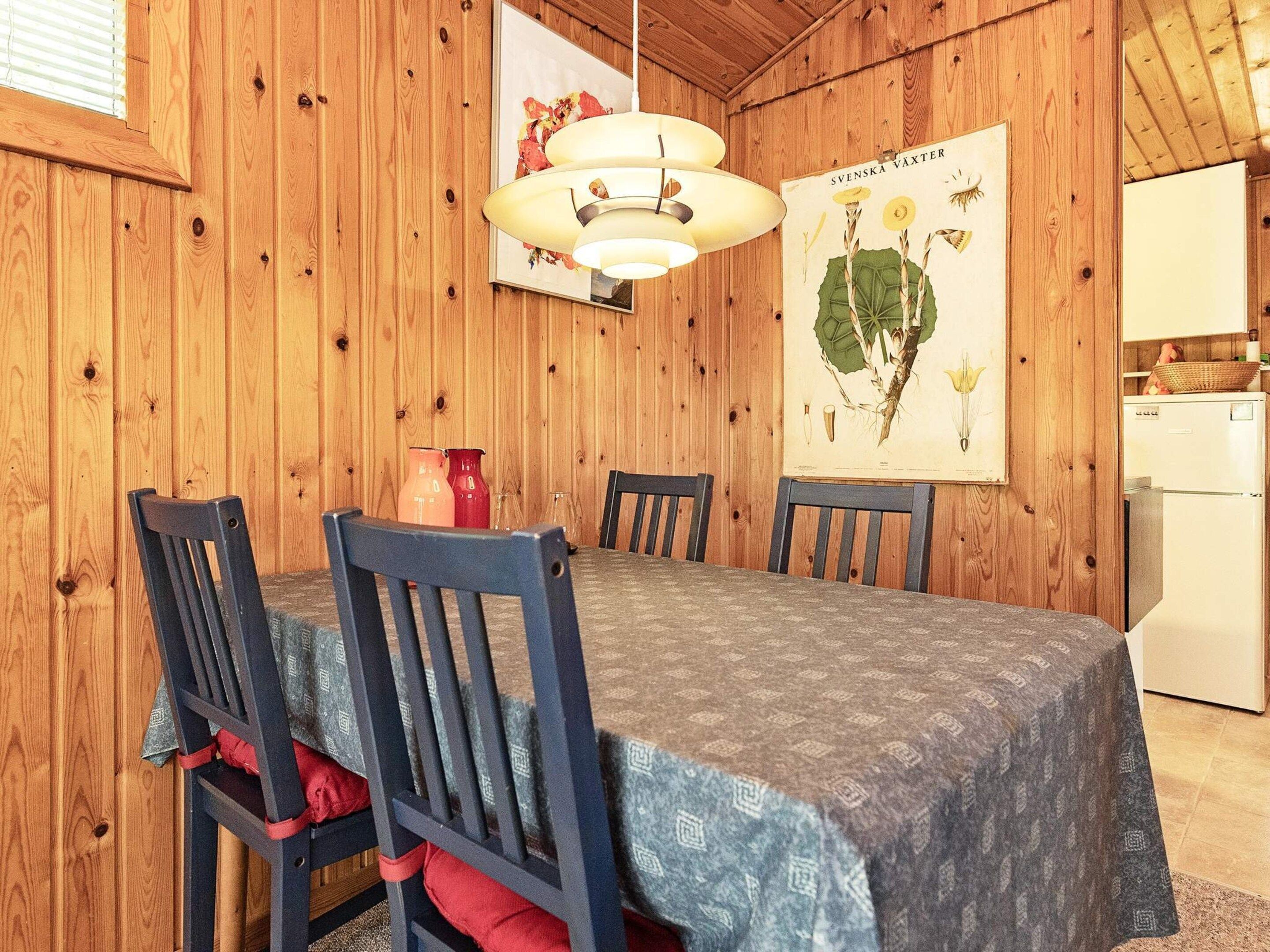 A wooden interior with a dining table, chairs, a hanging light fixture, and a framed botanical illustration on the wall.