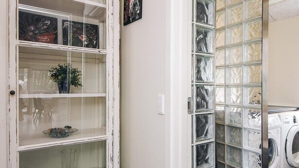 Una sala de bugaderia amb una rentadora, un moble d'estanteries amb frontissa de vidre i objectes decoratius, i una porta amb marc blanc.