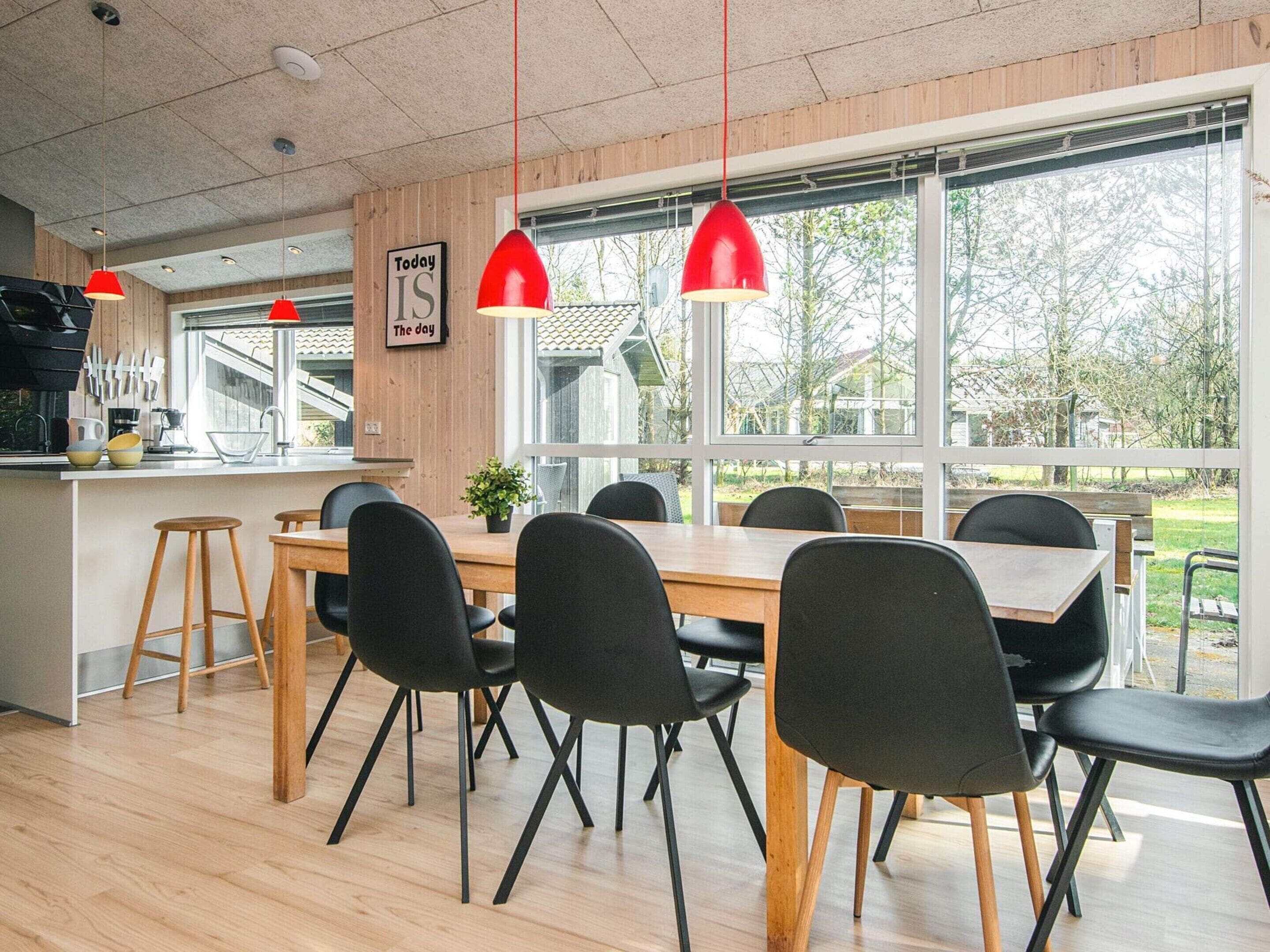 Een moderne eetruimte met een houten tafel en zwarte stoelen, grote ramen met uitzicht op groen en rode hanglampen.
