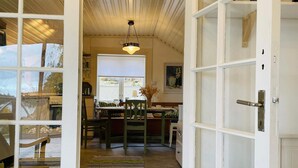 A cozy room with a wooden ceiling, a dining table with chairs, a hanging light fixture, and a view through a window.