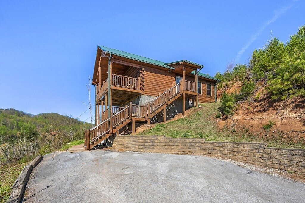 Una cabaña de troncos de dos pisos con un balcón y escaleras, rodeada por un paisaje montañoso.