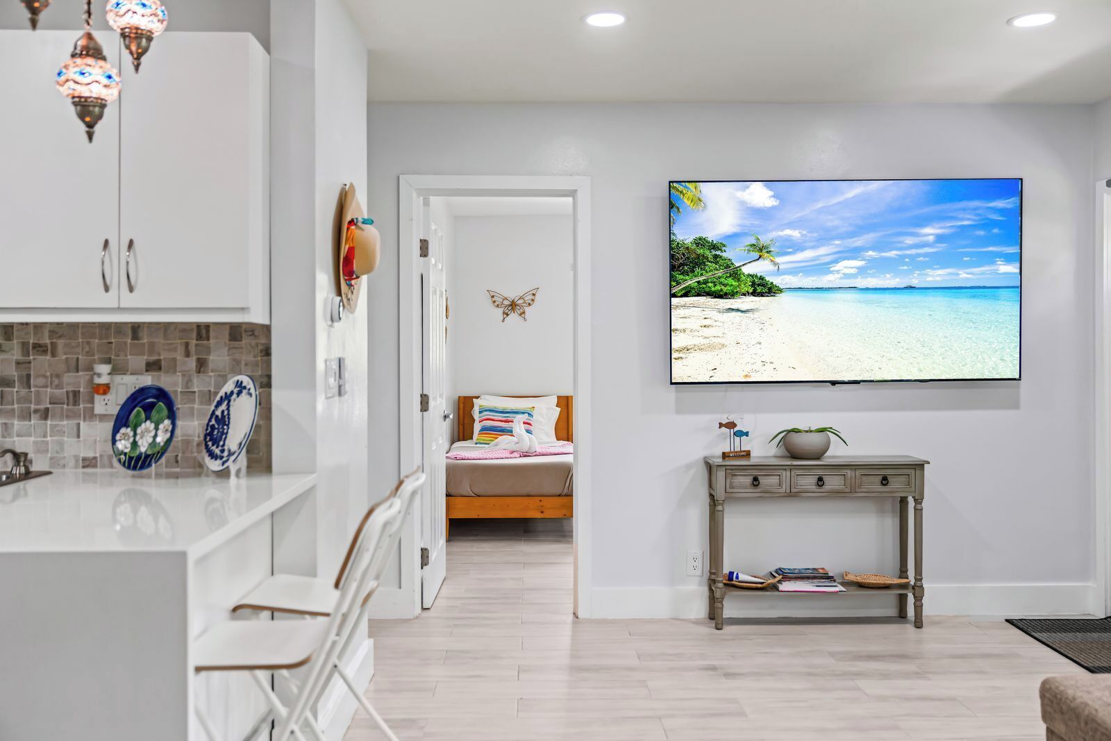 A modern hotel room with a kitchen area, a bed with a view of a beach, and a flat-screen TV on the wall.