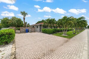 A paved driveway leading to a building with a tiled roof, surrounded by greenery and palm trees.