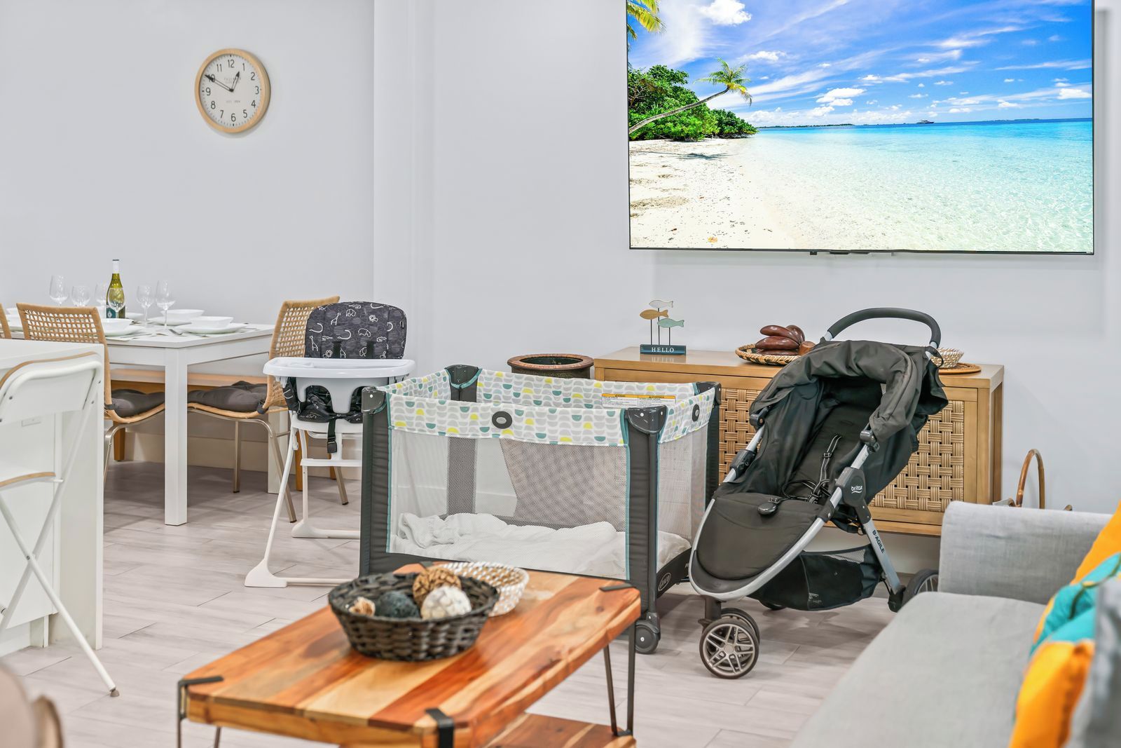A room with a dining table, chairs, a baby playpen, a stroller, and a large window with a beach view.