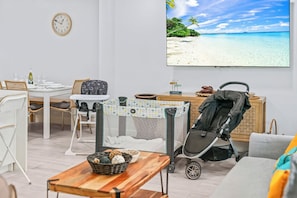 A room with a dining table, chairs, a baby playpen, a stroller, and a large window with a beach view.
