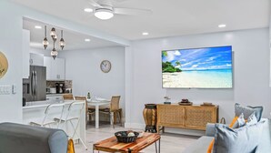 Una sala de estar moderna con un televisor de pantalla plana mostrando una escena de playa, un comedor con un reloj y una cocina con un refrigerador.